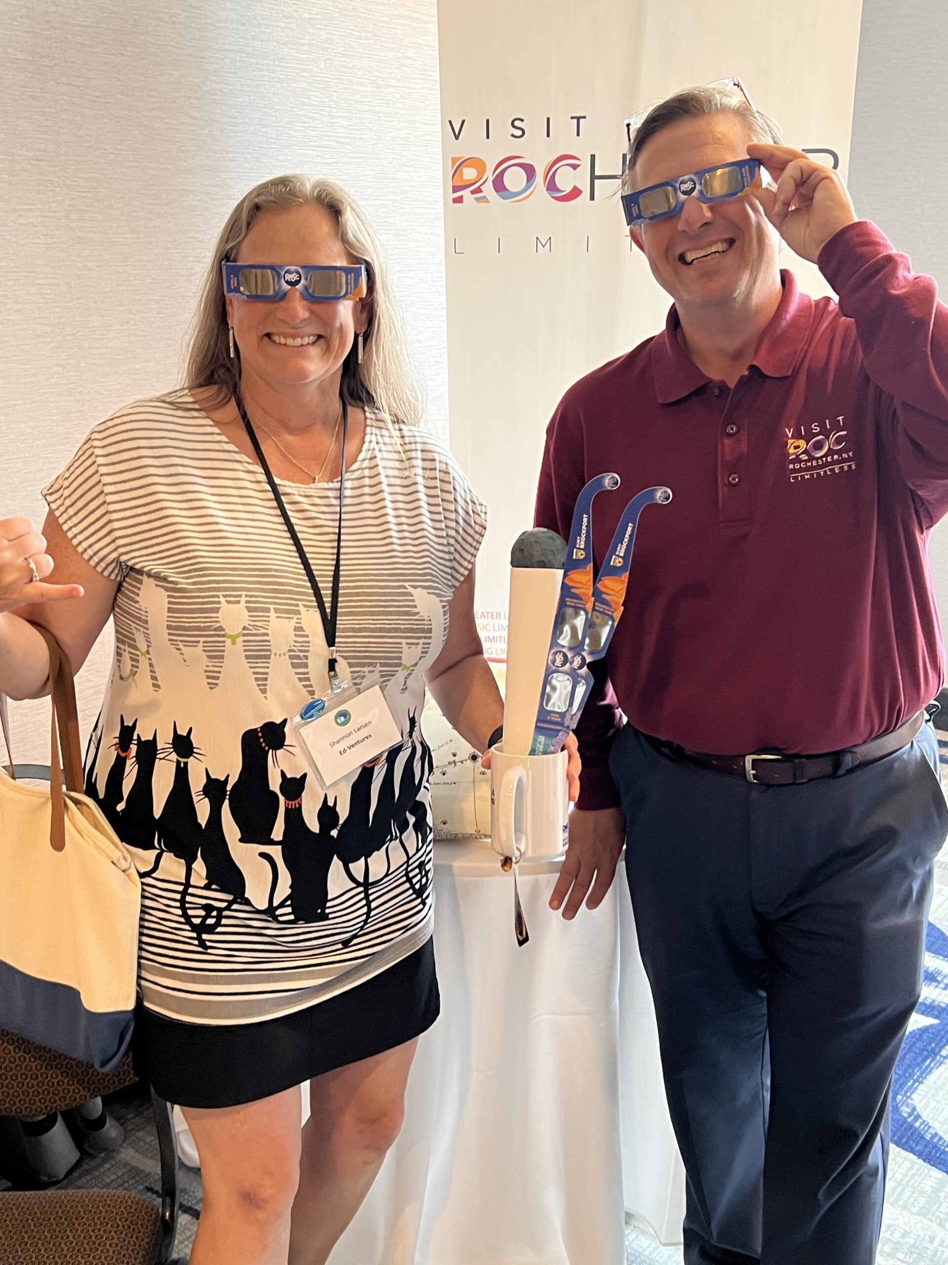 a man and a woman stand in front of a white background, wearing special eclipse glasses branded for Visit Rochester